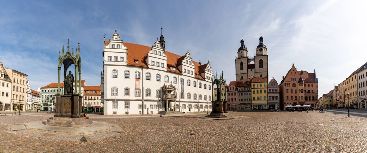 A cobblestone square with historic buildings, a church with twin spires, and a statue in the center.