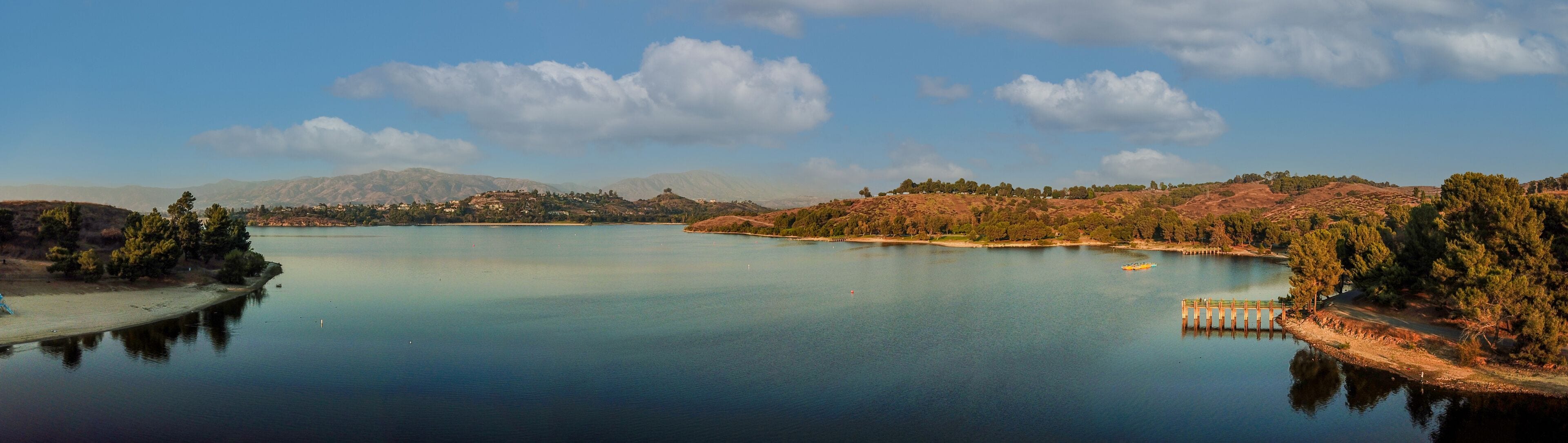 a breathtaking aerial panoramic shot of the still blue waters and lush green trees and majestic mountain ranges at Frank G Bonelli Regional Park at Puddingstone Lake in San Dimas California