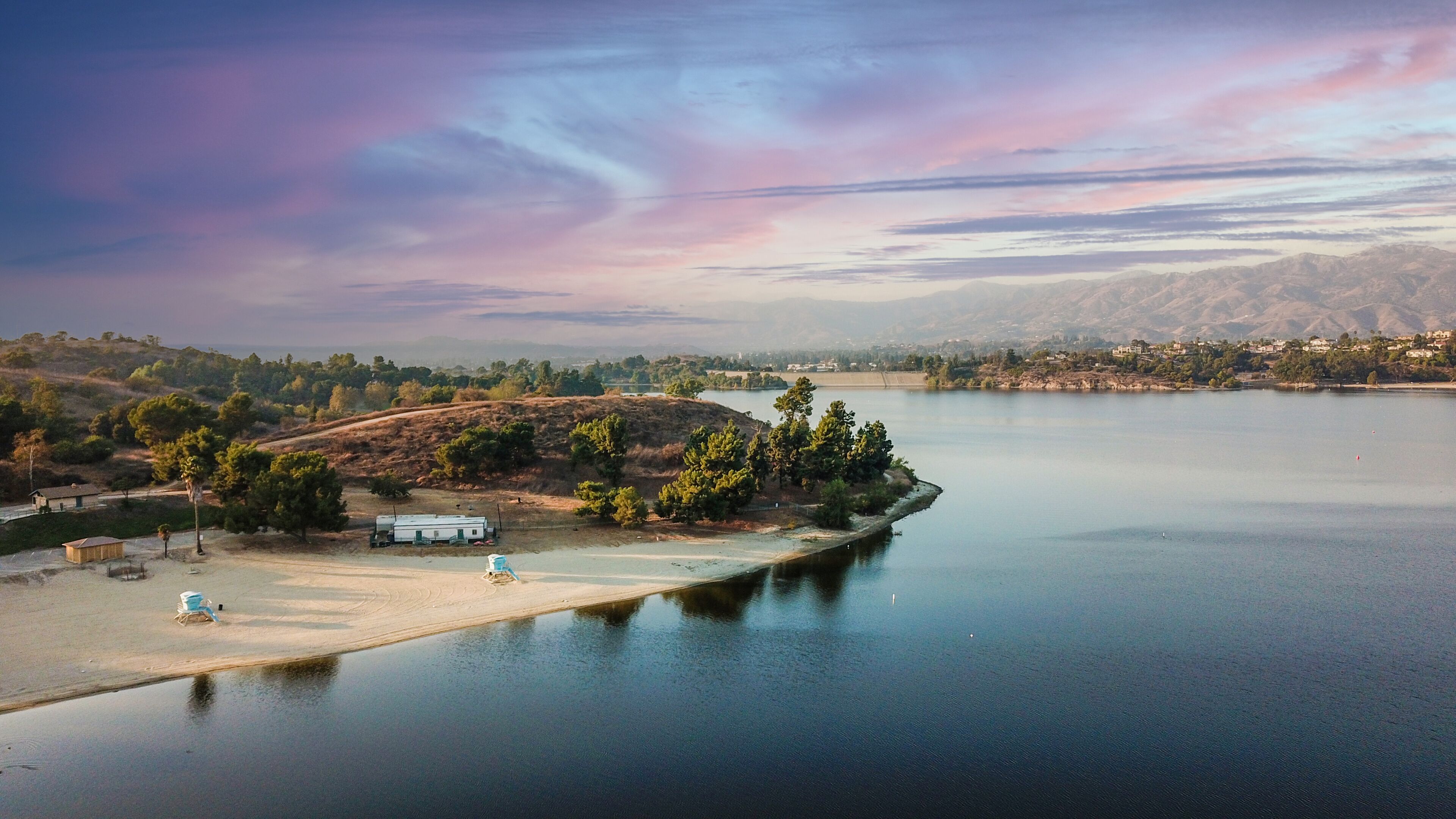 a stunning aerial shot of the still blue waters, the pier, the lush green trees and majestic mountain ranges at Puddingstone Lake in San Dimas California USA