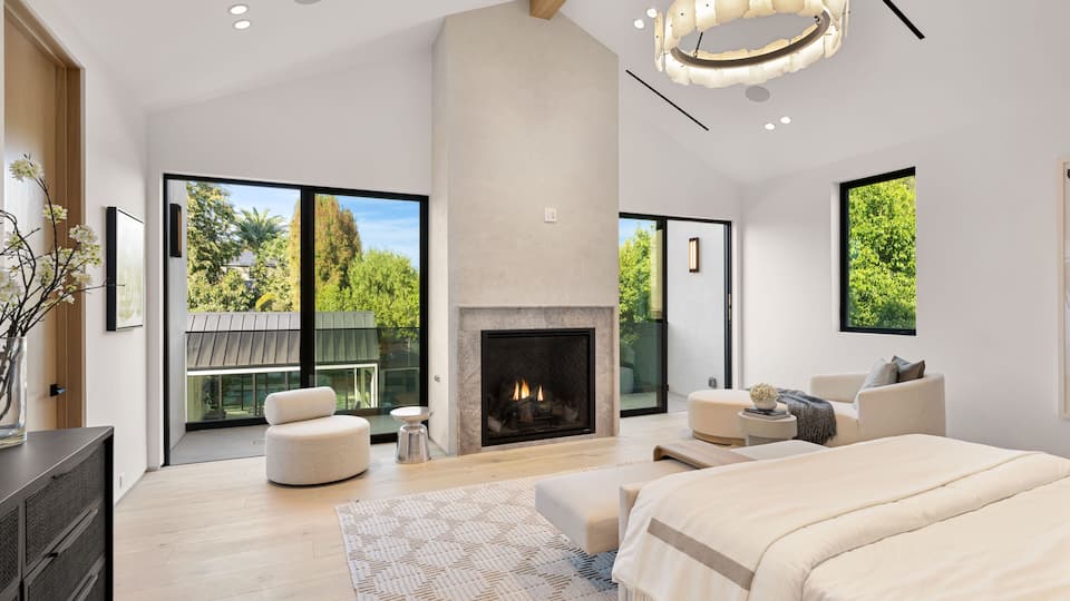 A modern bedroom with a fireplace, large windows, and a view of greenery.