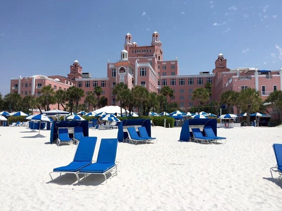 A beach with blue lounge chairs and umbrellas, a large pink building with towers, and palm trees.