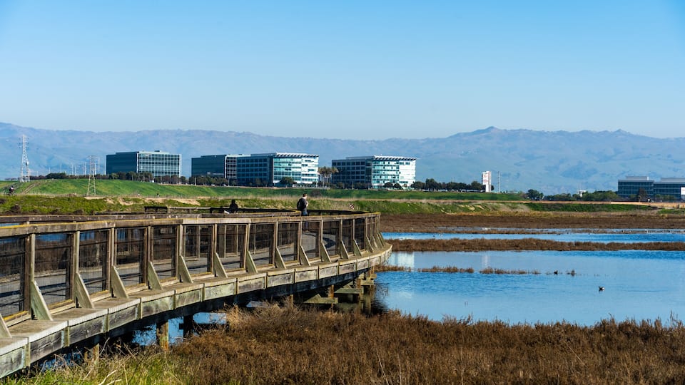 A wooden bridge over a body of water with a cityscape and mountains in the background.
