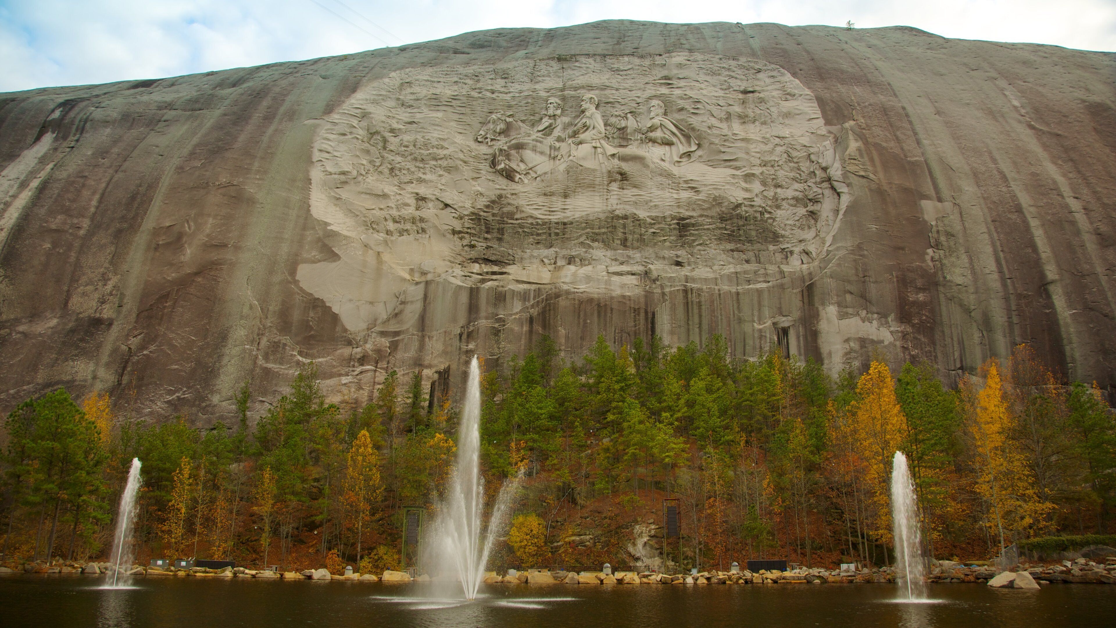 A large rock formation with a carved face, a lake with fountains, and autumn-colored trees.
