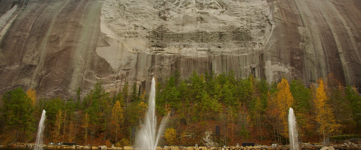 A large rock formation with a carved face, a lake with fountains, and autumn-colored trees.