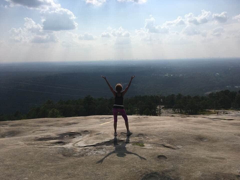 A person standing on a rocky outcrop with arms raised, overlooking a vast landscape with a distant city skyline.