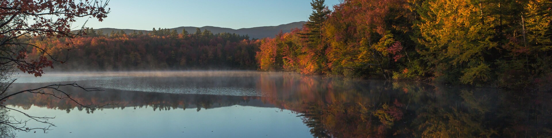 Un lago tranquilo que refleja el follaje otoñal y un bosque brumoso.