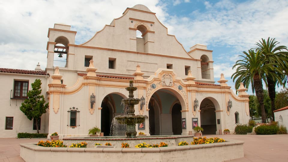 A historic building with a fountain and arches, surrounded by a plaza with flowers and palm trees.