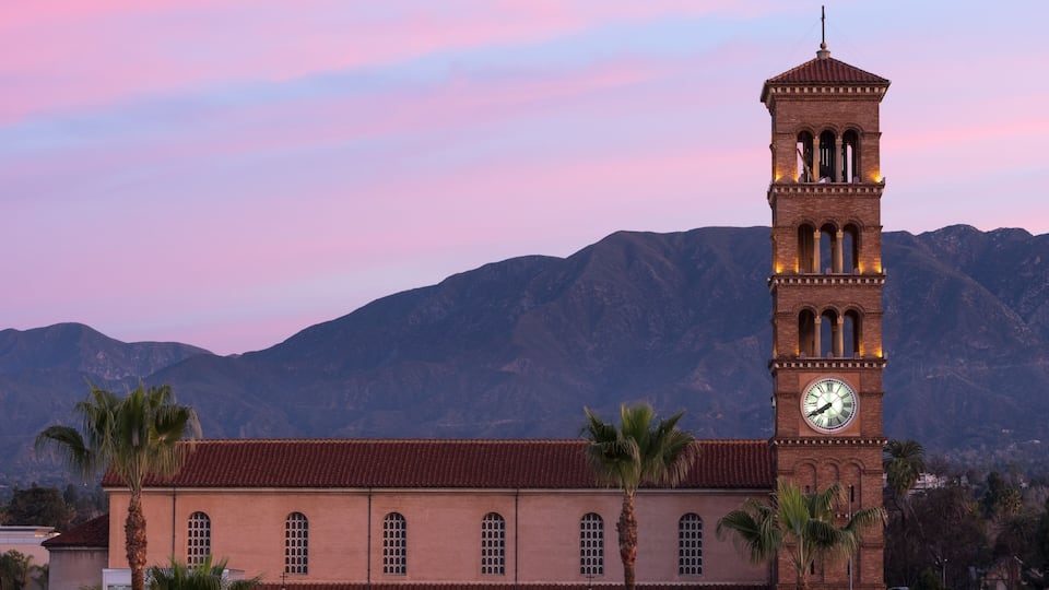 A clock tower with a red-tiled roof, palm trees, and mountains in the background.