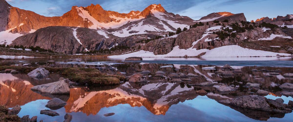 A mountain landscape with a lake reflecting the peaks, surrounded by snow patches and rocky terrain.