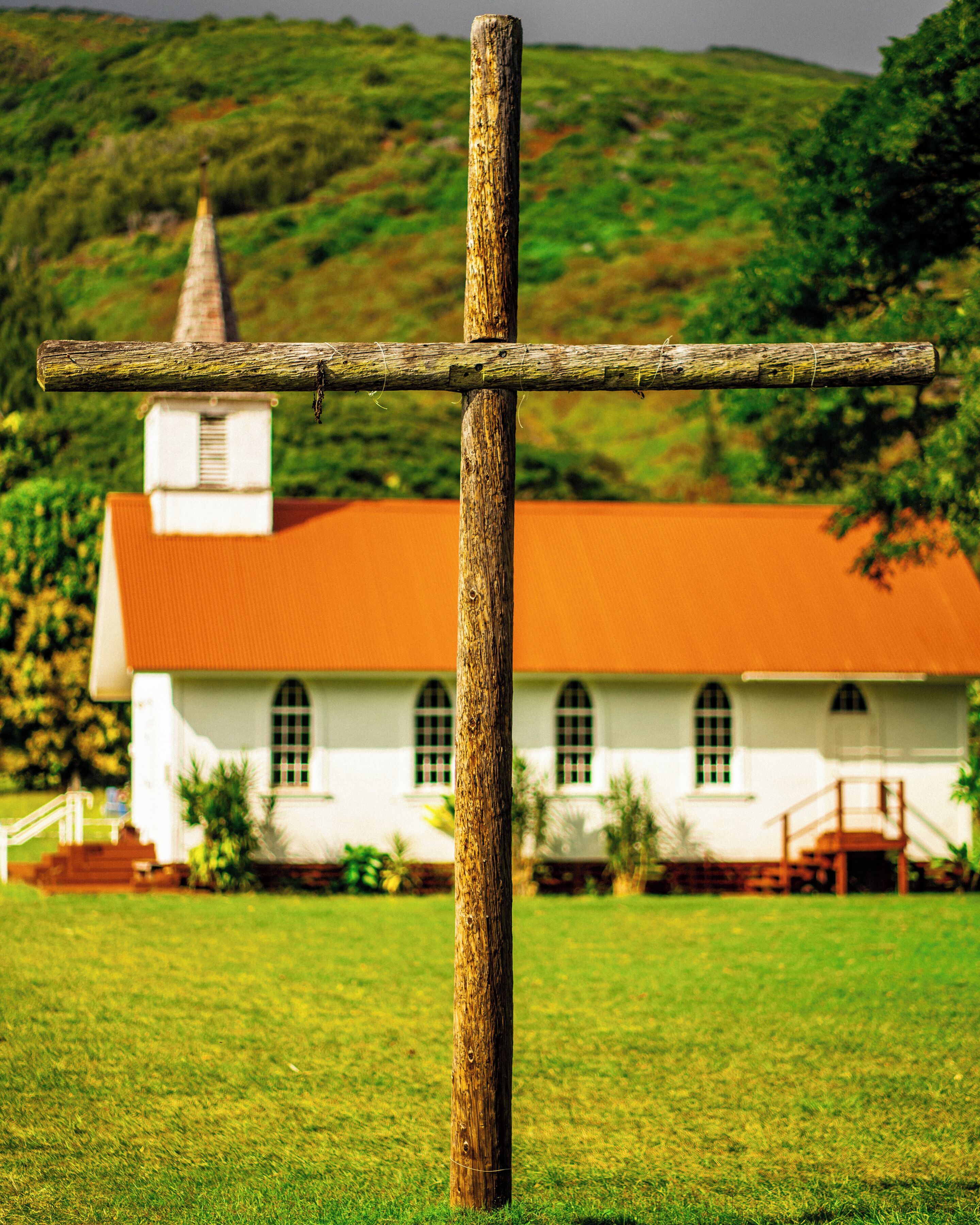 A wooden cross in a grassy field with a white building with a red roof in the background.