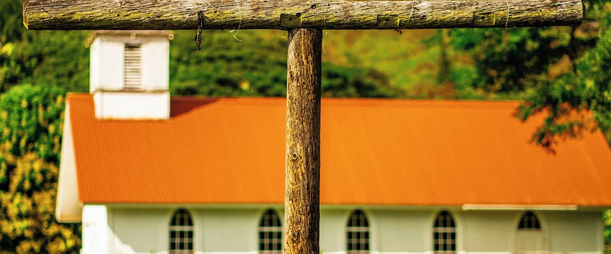 A wooden cross in a grassy field with a white building with a red roof in the background.