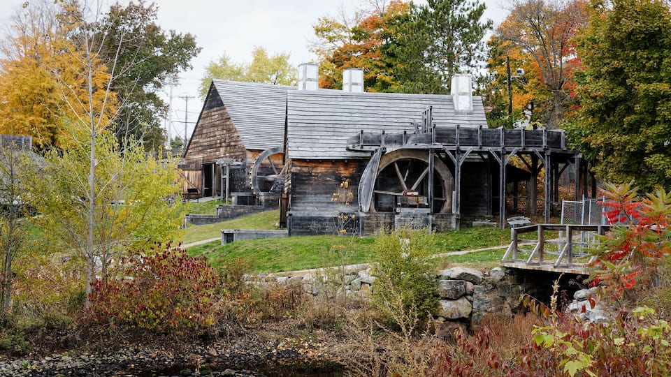 A rustic log cabin with a water wheel and a stone bridge in a wooded area.