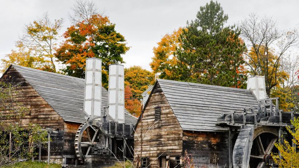 A rustic watermill with wooden structures and large water wheels, surrounded by autumn foliage and a grassy foreground.