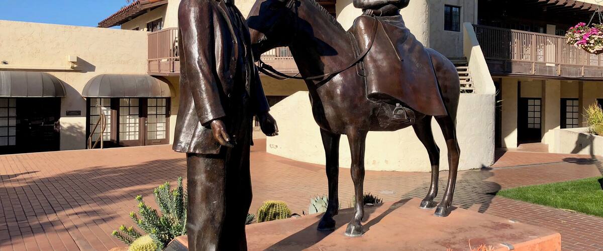 A bronze statue of a man with a horse in a courtyard with buildings and plants.