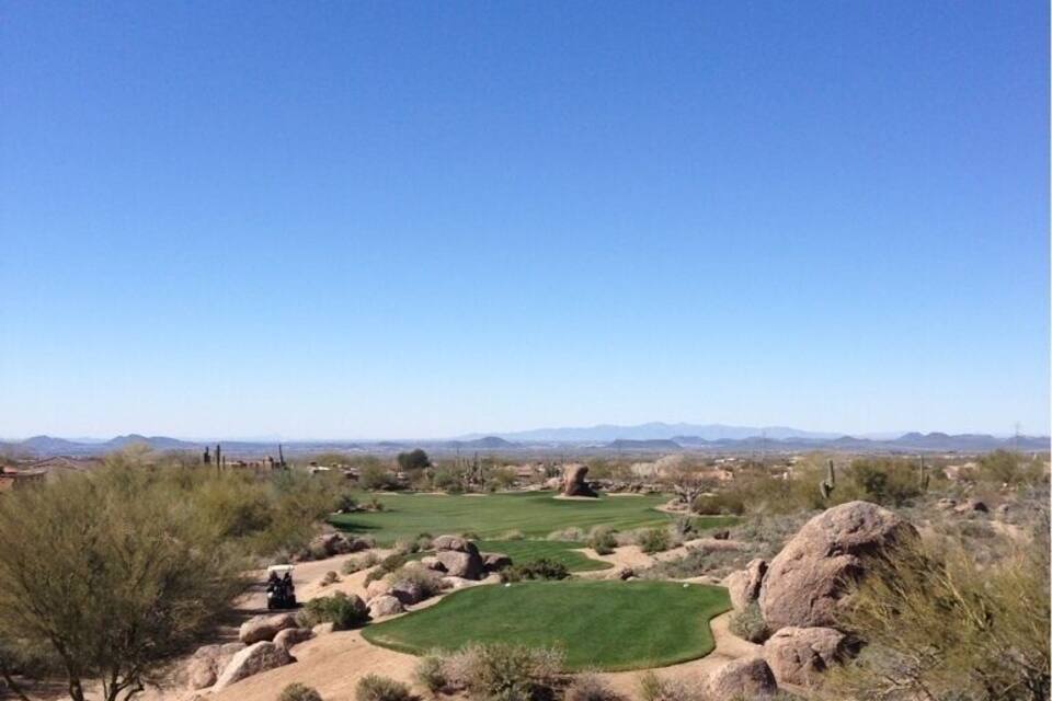 A golf course with a clear blue sky, surrounded by desert vegetation and distant mountains.