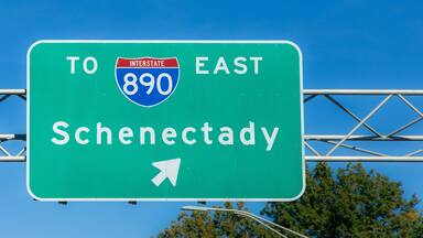 A green highway sign directing drivers to Interstate 890 East toward Schenectady, set against a clear blue sky, indicating travel directions and regional navigation.