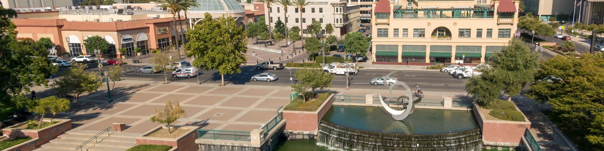 Uma praça urbana com uma fonte central, edifícios ao redor e céu claro.