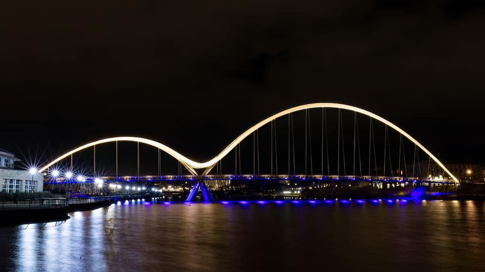 A nighttime view of a large, illuminated bridge with a distinctive arch design over a body of water.