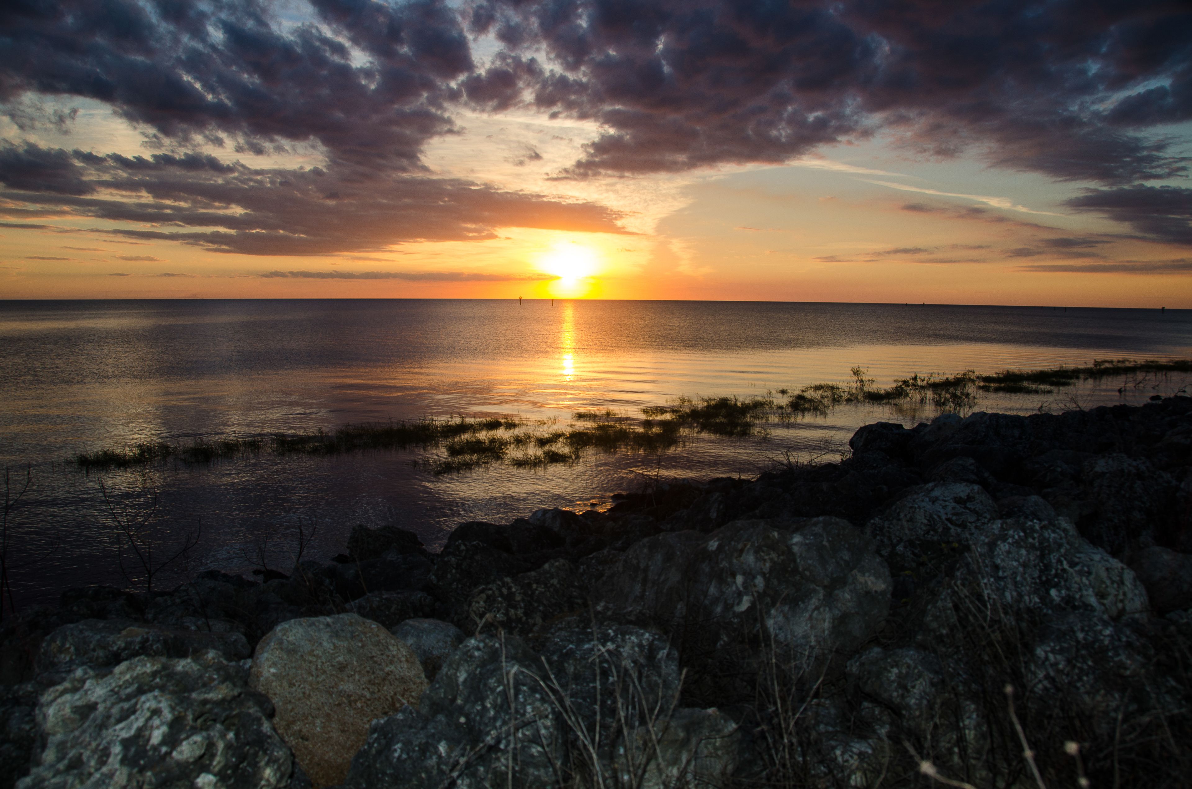 A sunset over a calm sea with a rocky shoreline in the foreground.