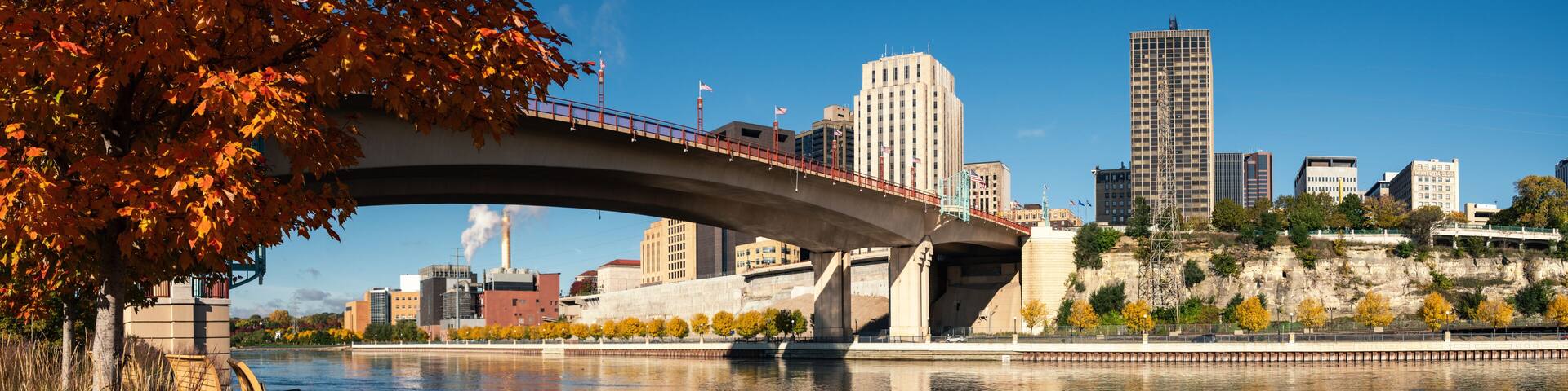 A bridge over a river with autumn leaves, a city skyline, and a statue.