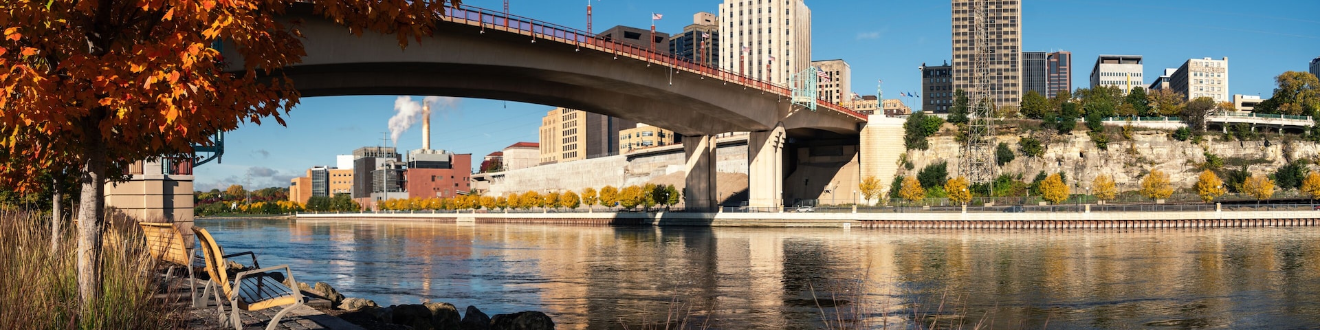 A bridge over a river with autumn leaves, a city skyline, and a statue.