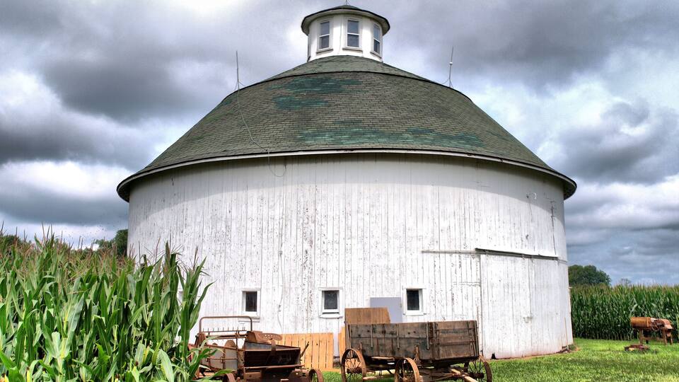 A white, round barn with a green roof, surrounded by cornfields and a wooden cart.