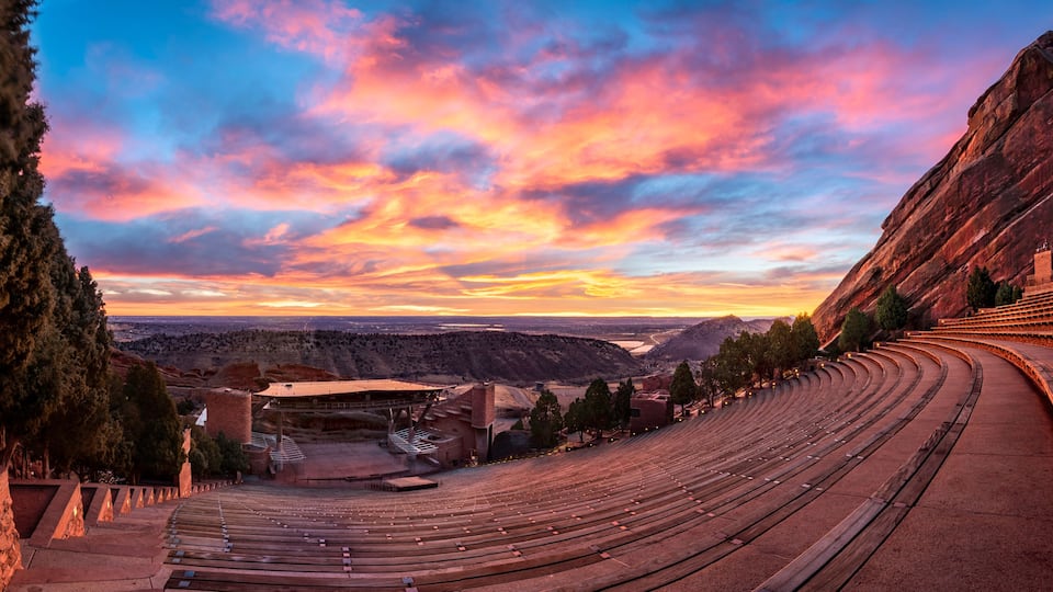 A large outdoor amphitheater with tiered seating, set against a backdrop of a mountainous landscape and a colorful sunset sky.