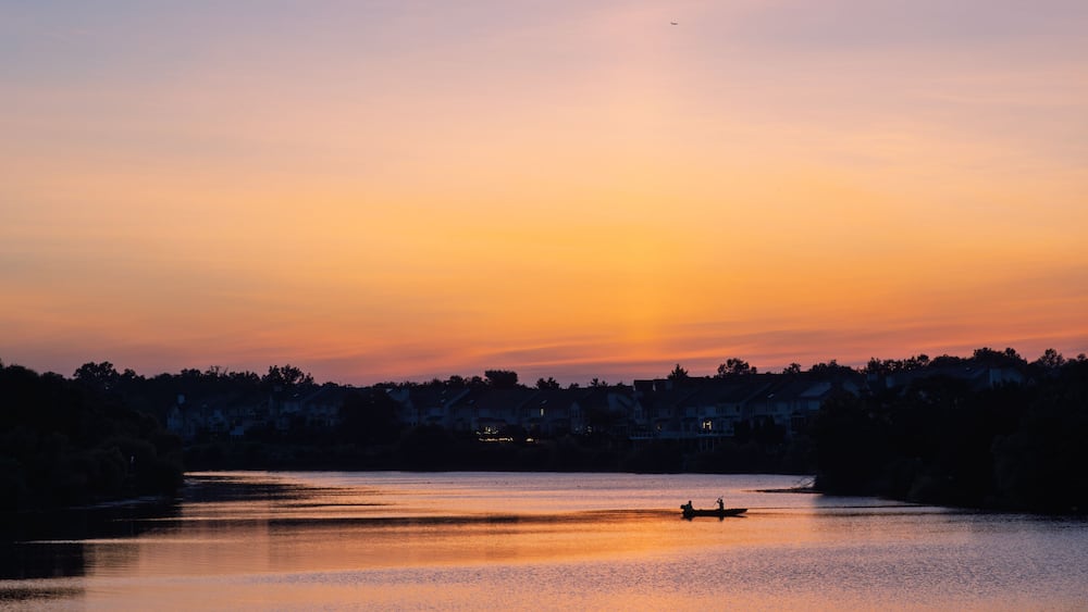 A sunset over a calm lake with a silhouette of a boat and houses in the background.