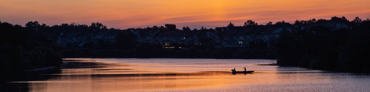 A sunset over a calm lake with a silhouette of a boat and houses in the background.