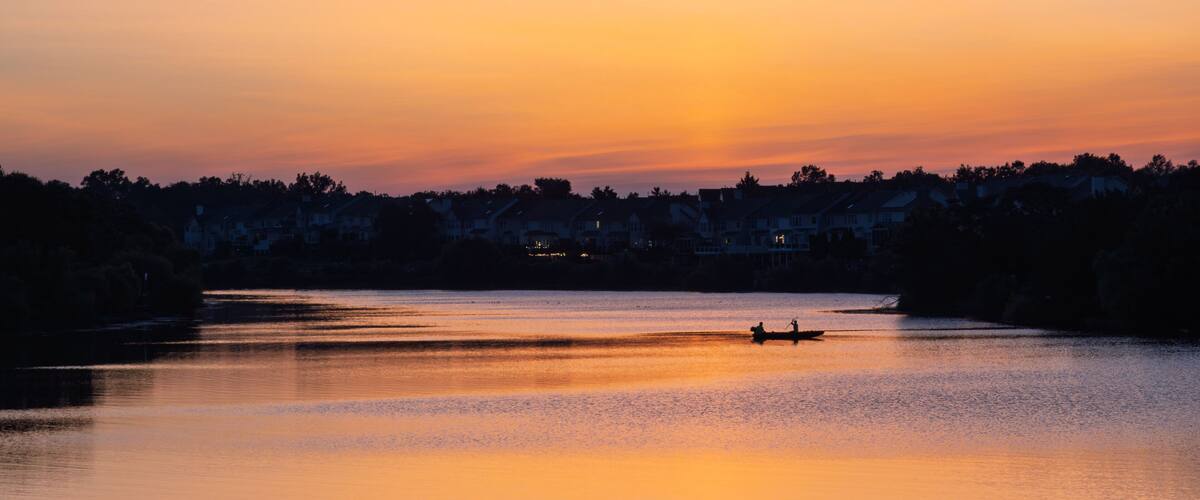 Un atardecer sobre un lago tranquilo con la silueta de un bote y casas al fondo.