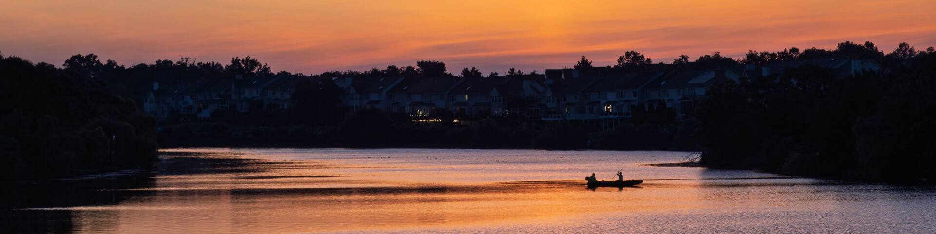 A sunset over a calm lake with a silhouette of a boat and houses in the background.
