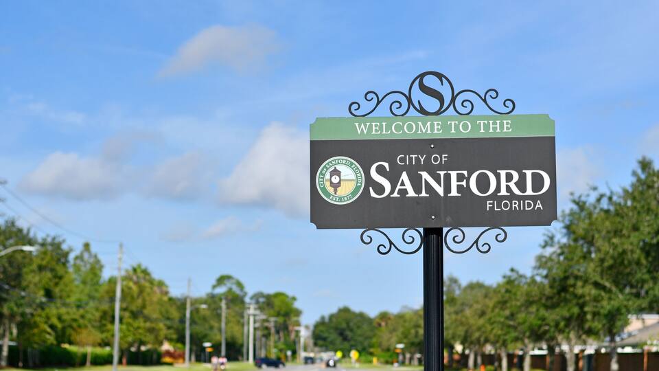A welcome sign for the City of Sanford, Florida, with a clear sky and a road lined with trees.