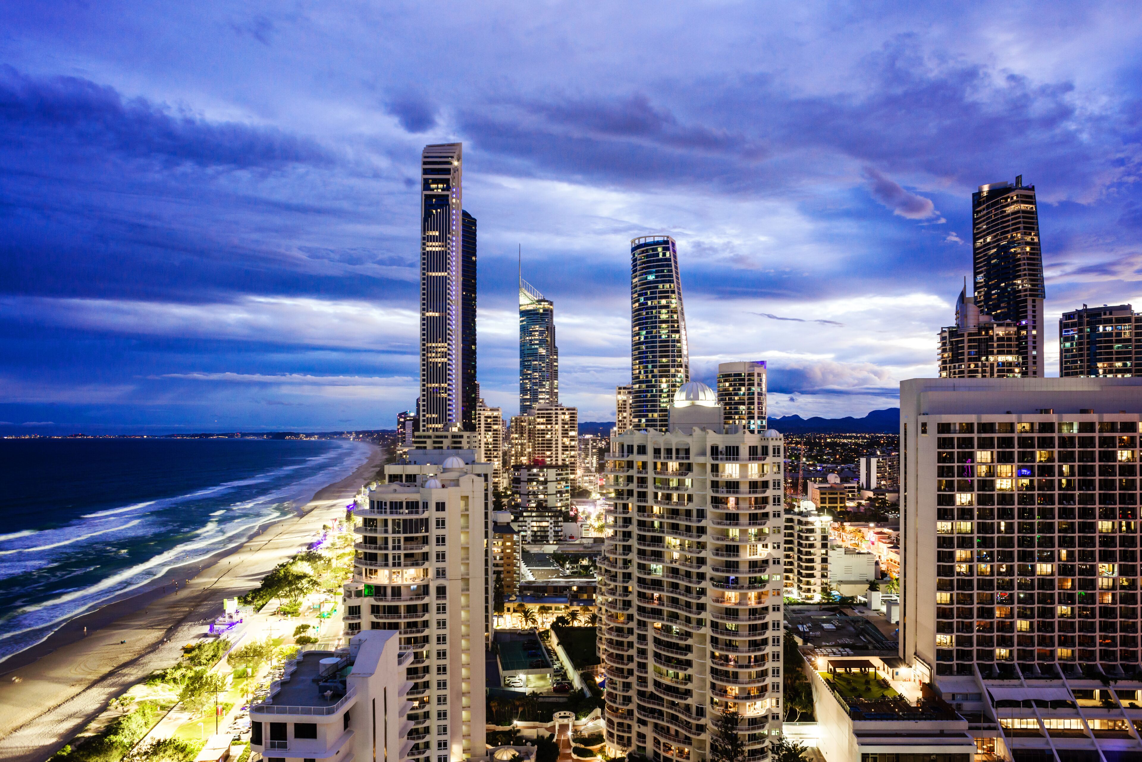 Paisagem costeira com edifícios altos, uma praia e montanhas ao fundo.