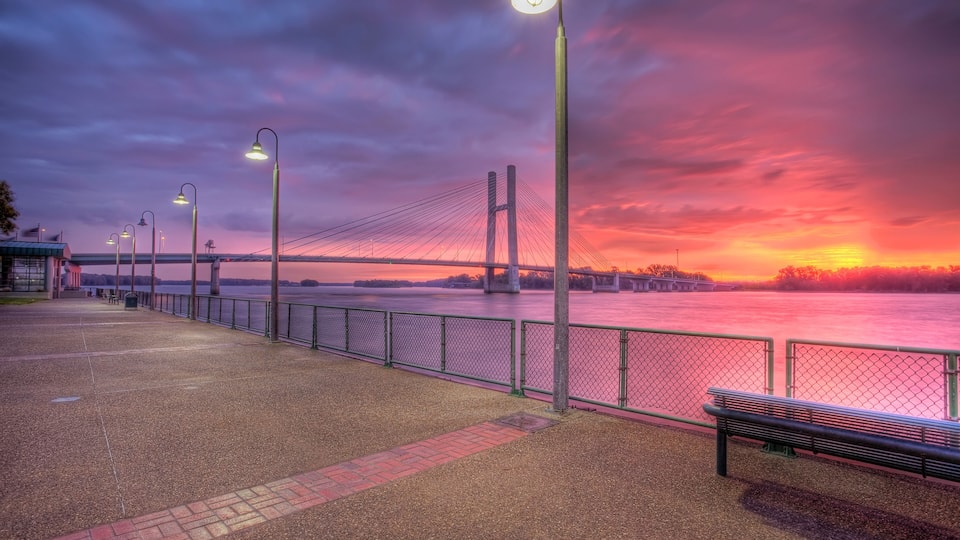 A waterfront promenade with street lamps, a bench, and a suspension bridge in the background.