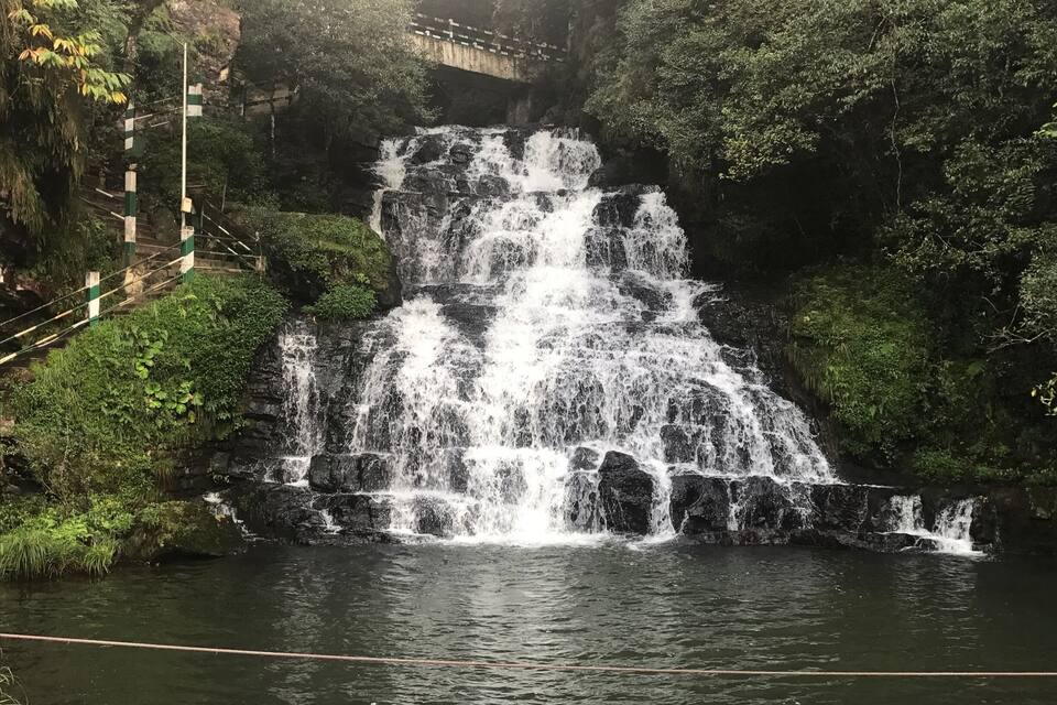Uma cascata em vários níveis deságua em uma piscina, cercada por vegetação exuberante e com uma ponte ao fundo.