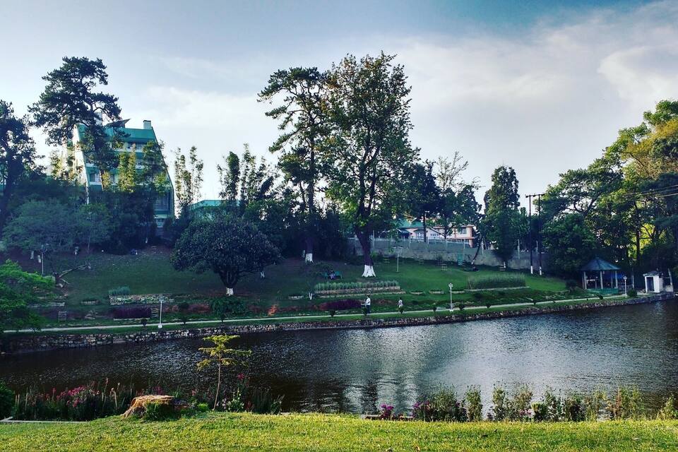 Um parque com lago, árvores, um coreto e um edifício ao fundo.