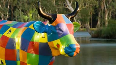 A colorful, rainbow-patterned cow sculpture with a water body and trees in the background.