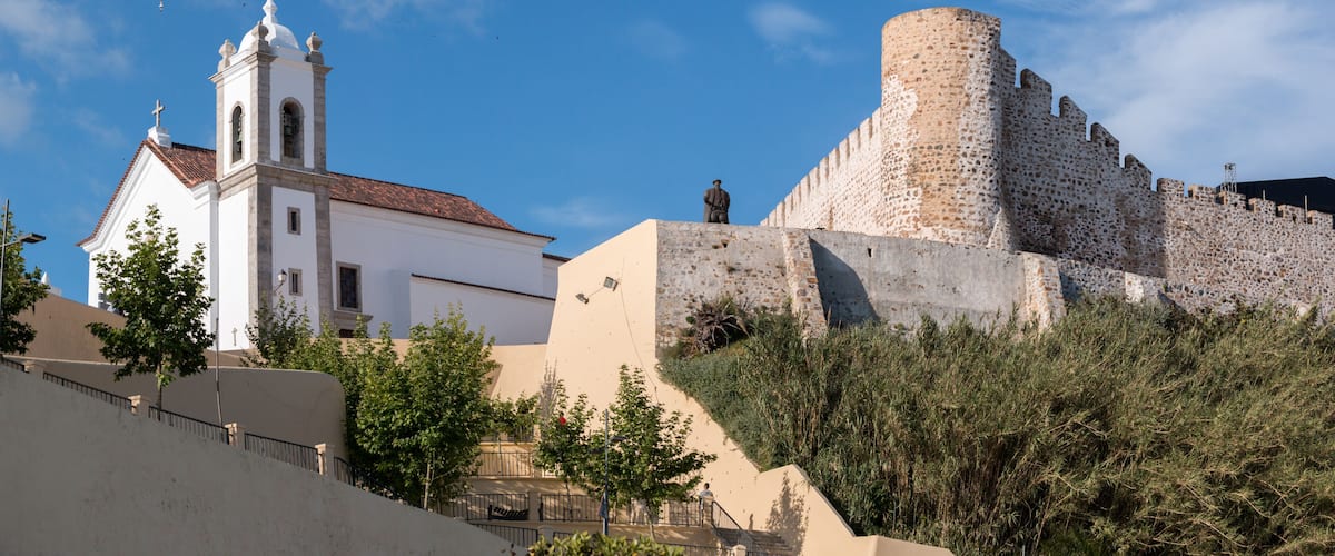 A white church with a bell tower and a large stone fortress with a round tower.