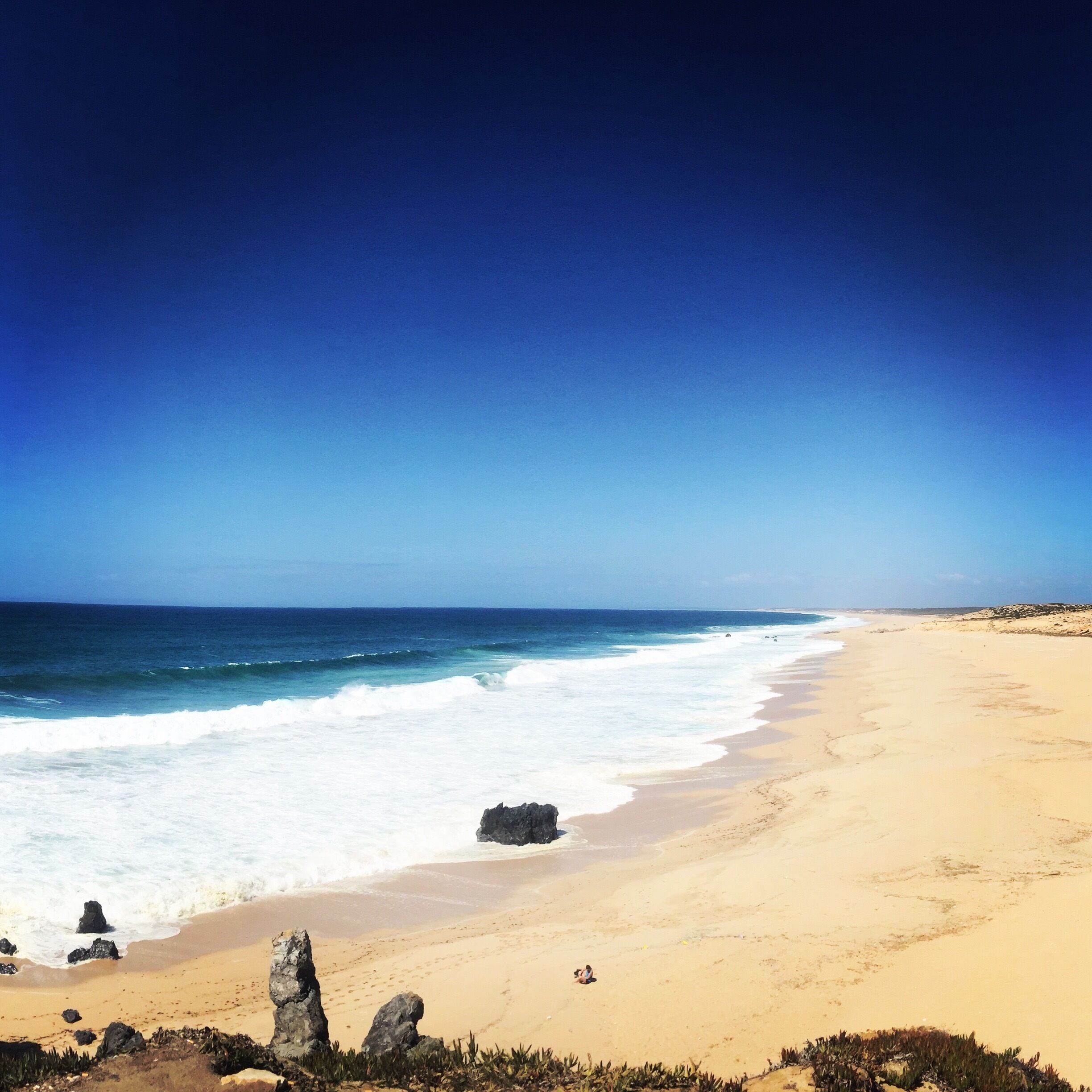 Uma praia arenosa com ondas quebrando na costa e um céu azul límpido.