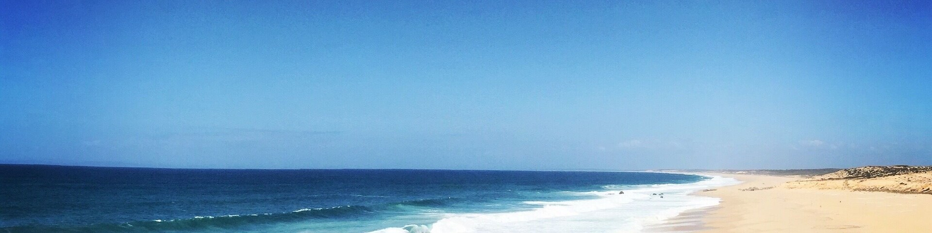 Uma praia arenosa com ondas quebrando na costa e um céu azul límpido.