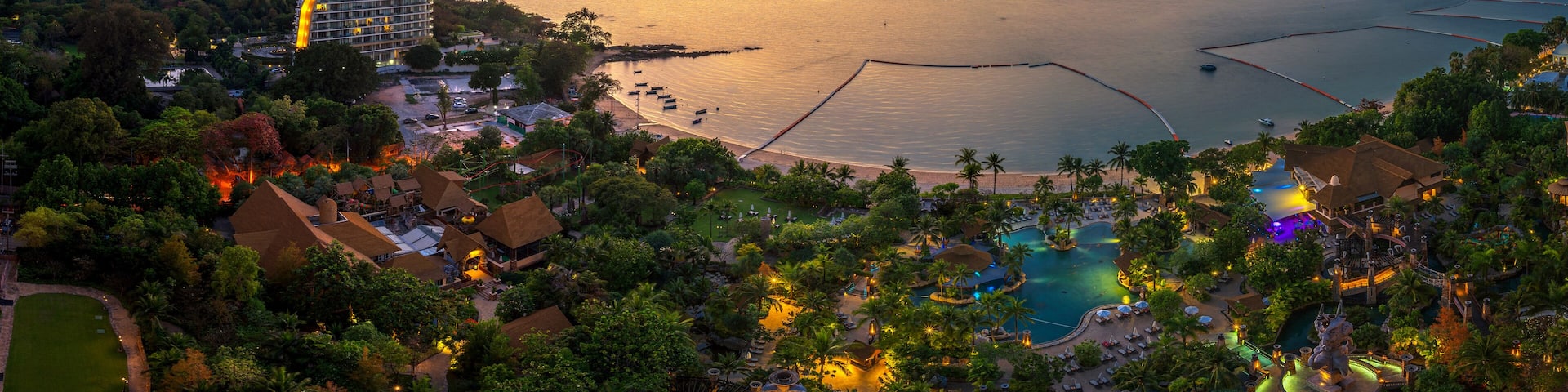 A coastal resort with a large pool, palm trees, and a sunset over the water.