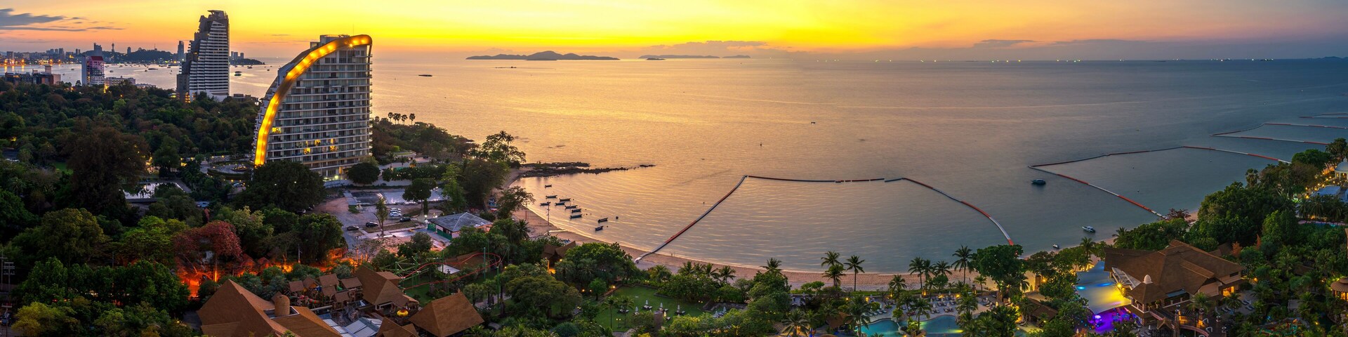 A coastal resort with a large pool, palm trees, and a sunset over the water.