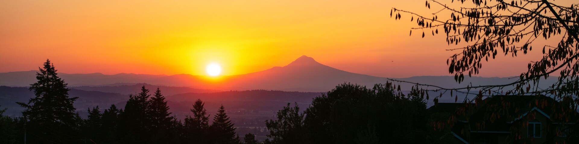 Solnedgang over et bjerglandskab med et hus i forgrunden.