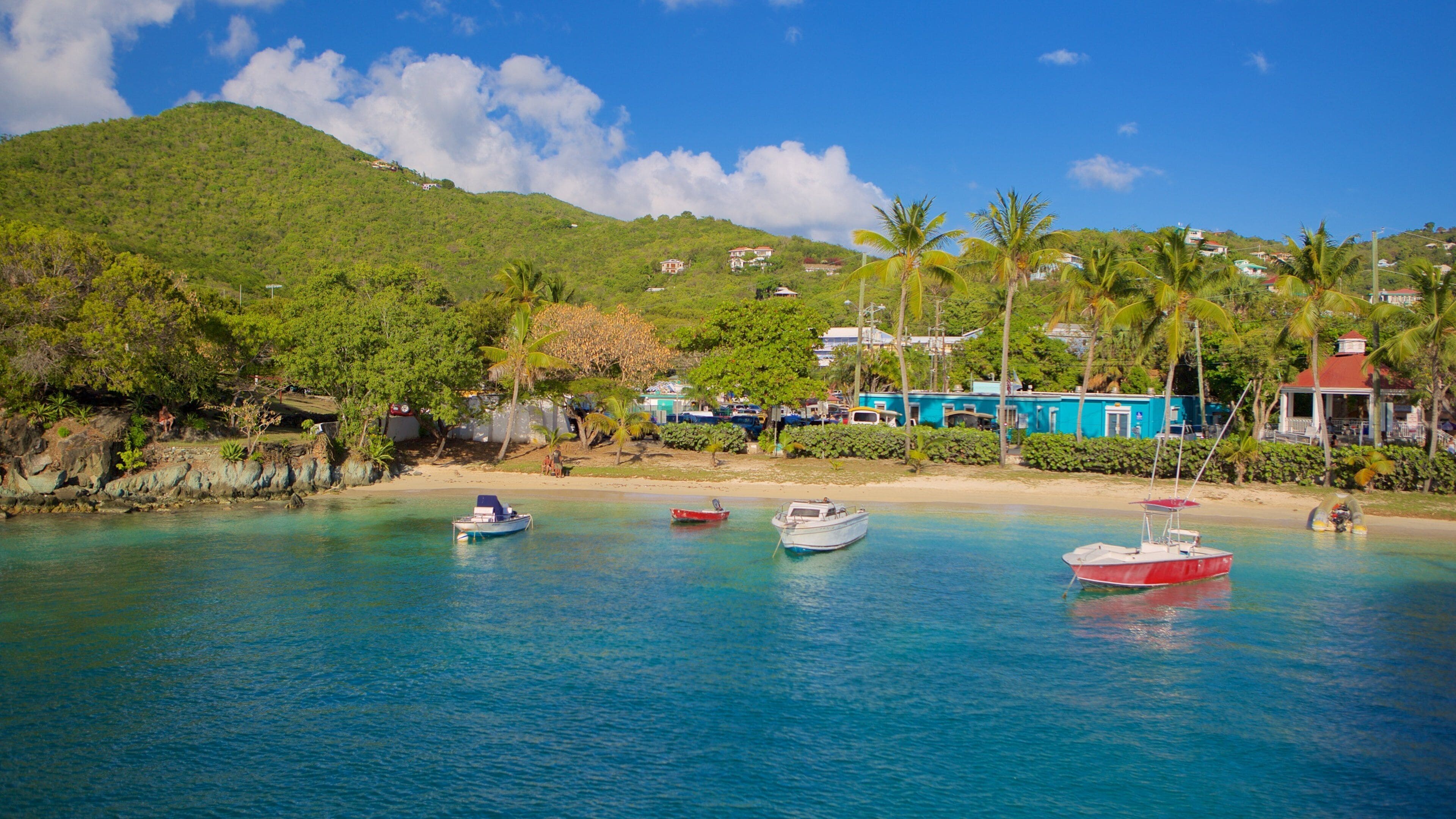 A tropical beach with clear turquoise waters, several boats anchored near the shore, and a backdrop of lush green hills and a blue building.