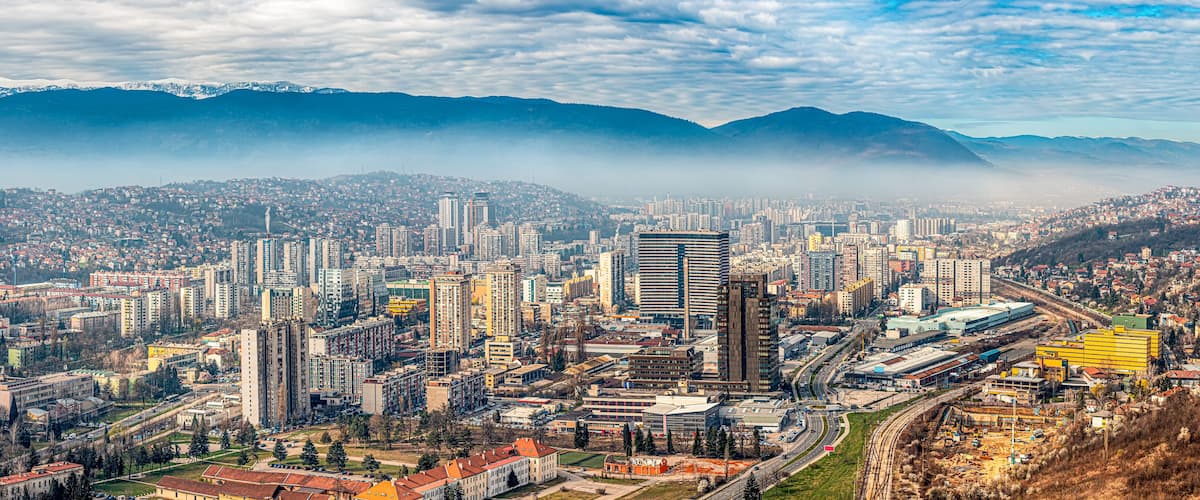 A cityscape with a prominent modern building, a dense urban area, and mountains in the background.