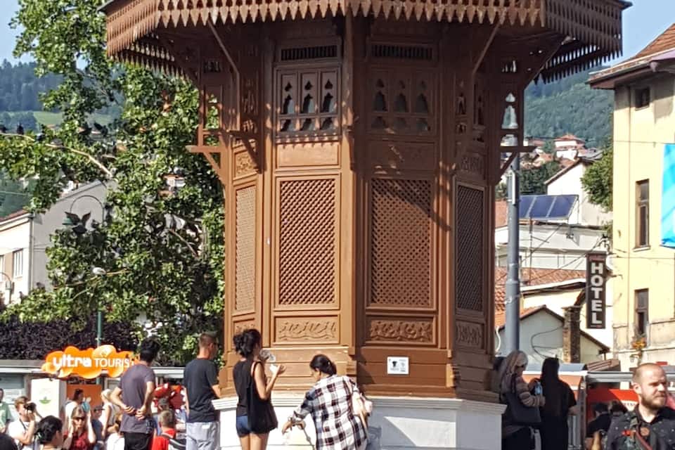 A traditional wooden pavilion with a green dome, surrounded by people and trees.
