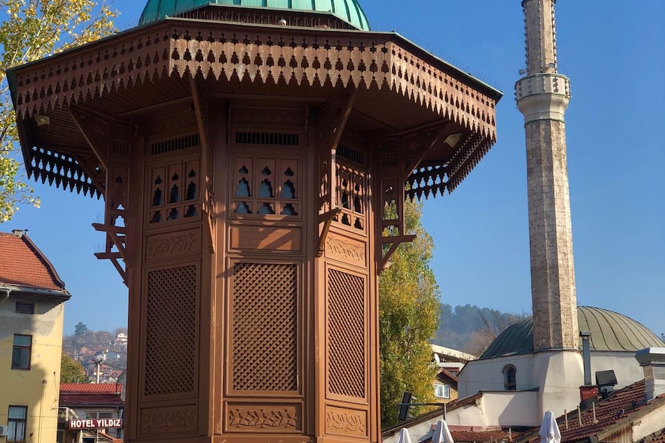 A traditional wooden pavilion with a green dome, a tall minaret, and a bustling street scene with cafes and shops.