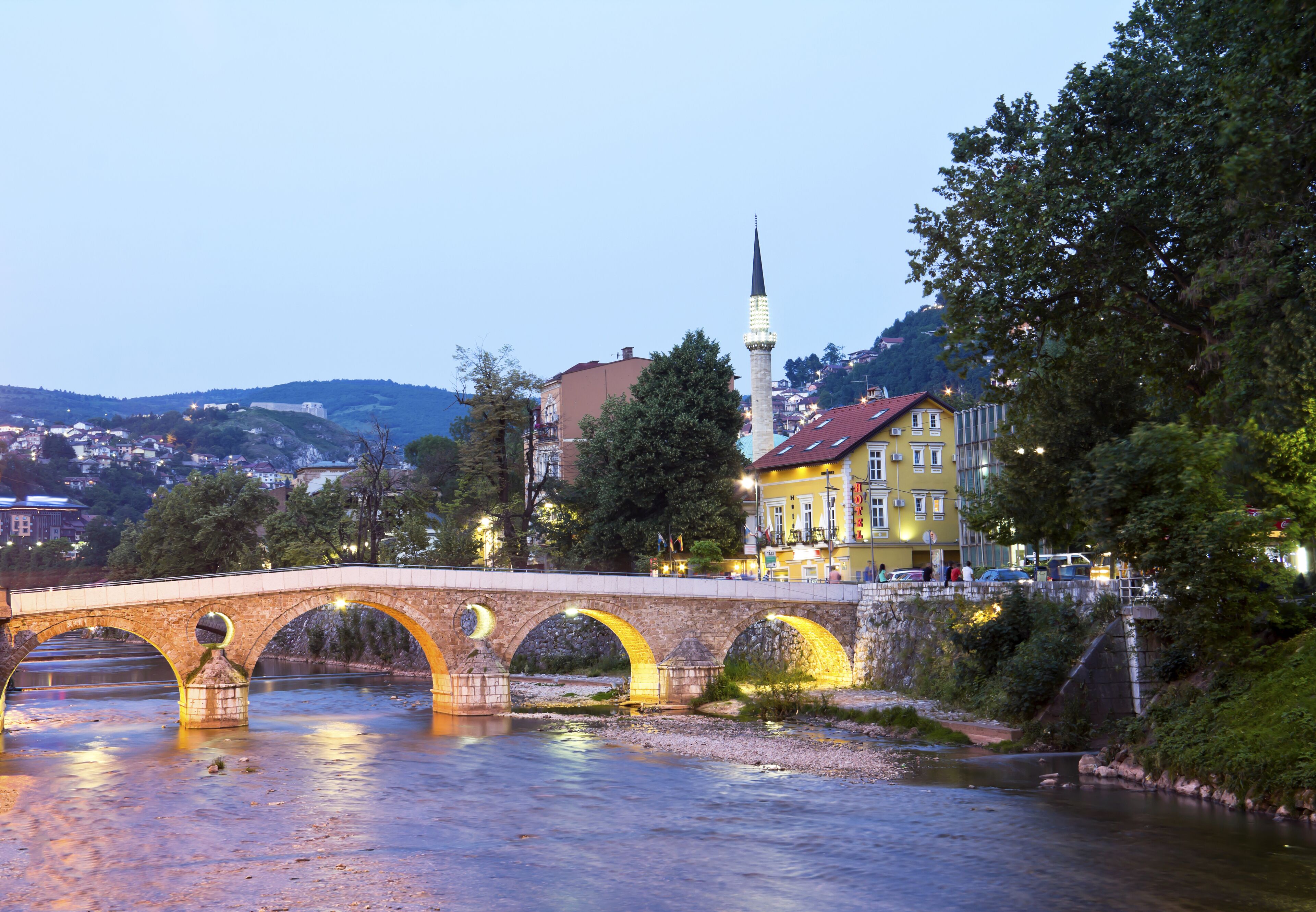 A stone bridge over a river, a mosque with a tall minaret, and a yellow building with a red roof.