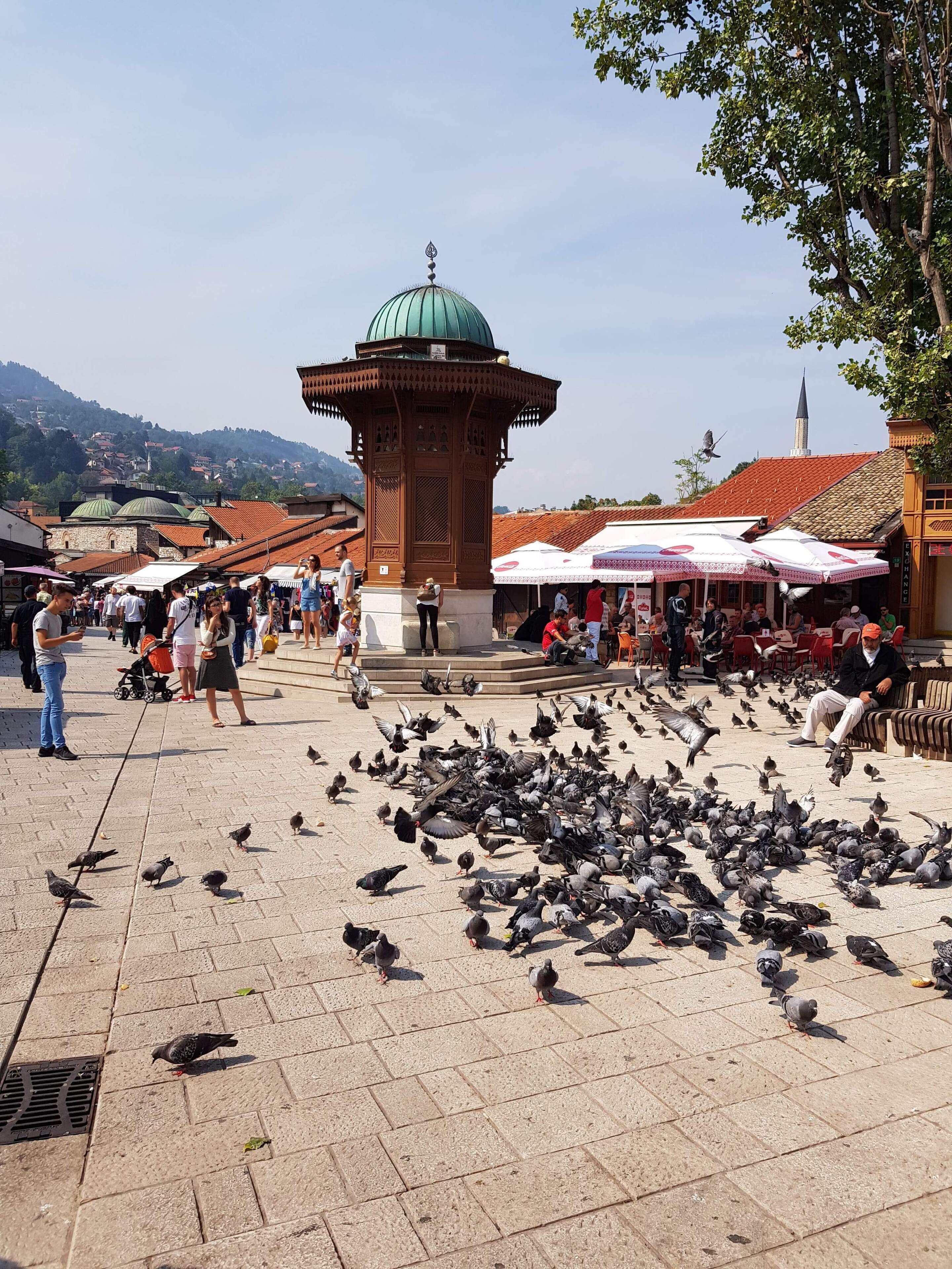 A bustling square with a central tower, surrounded by people and pigeons.