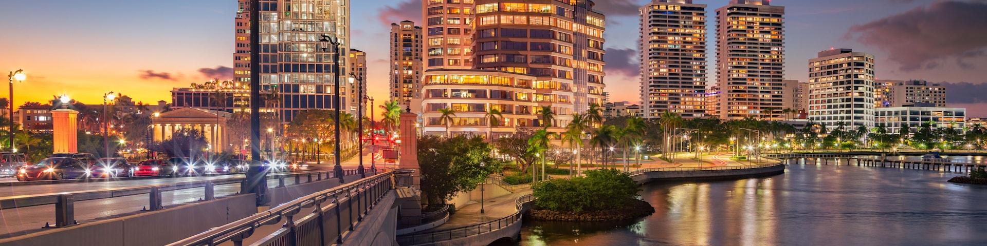 Horizonte urbano al atardecer con edificios iluminados, un puente y un río tranquilo que refleja las luces.